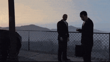 two men in suits standing on a balcony looking at their watch