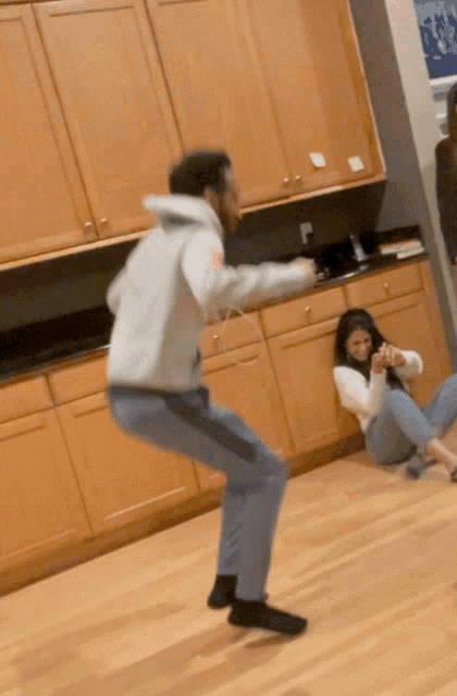 a man in a grey hoodie is jumping in the air while a woman sits on the floor