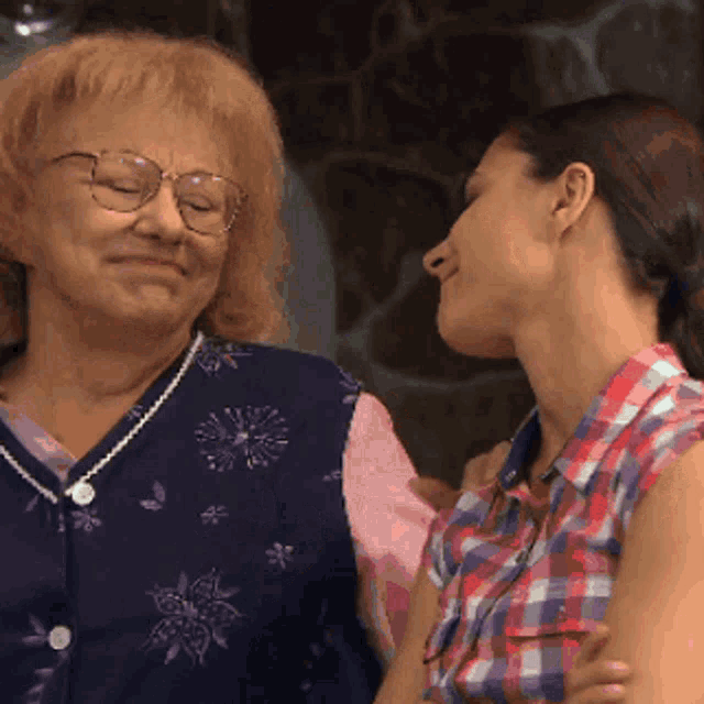 a woman wearing glasses and a plaid shirt kisses an older woman on the cheek
