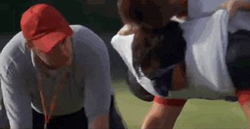 a man and a woman are doing push ups on a field . the man is wearing a red hat .