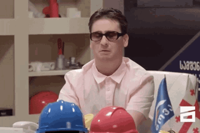 a man wearing sunglasses and a pink shirt sits at a table with hard hats and flags