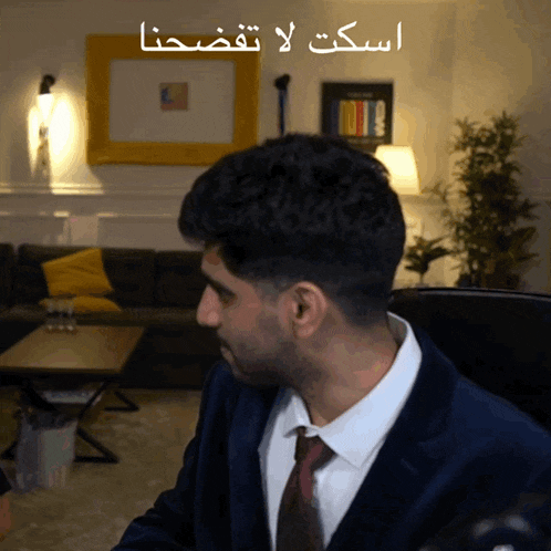 a man in a suit and tie is sitting in front of a couch with arabic writing above him