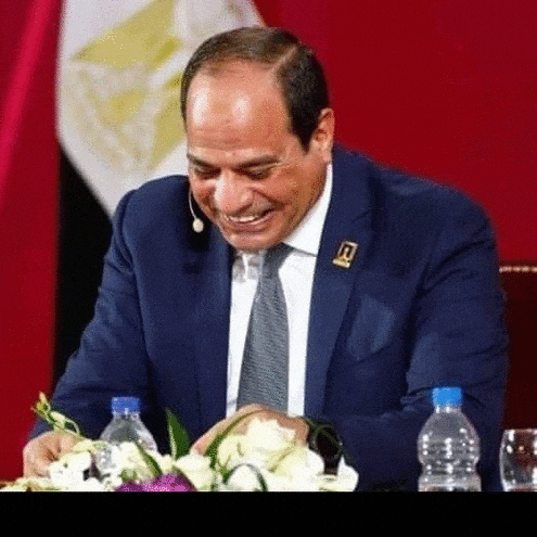 a man in a suit and tie is smiling while sitting at a table with flowers and bottles of water .