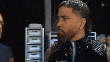 a man with a beard is wearing a black leather jacket and standing next to another man .