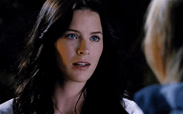 a woman with long dark hair and blue eyes looks at another woman in a dark room