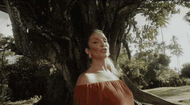 a woman in a red off the shoulder dress is standing in front of a tree