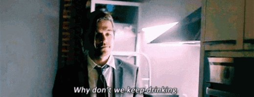 a man in a suit and tie is standing in a kitchen and says why don 't we keep drinking .