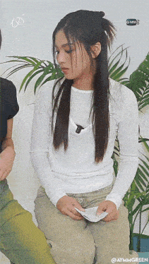 a woman in a white shirt is sitting in front of a palm tree with aymmgreen written in the corner