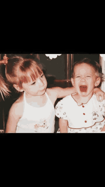 two little girls are sitting next to each other one is crying and the other is laughing