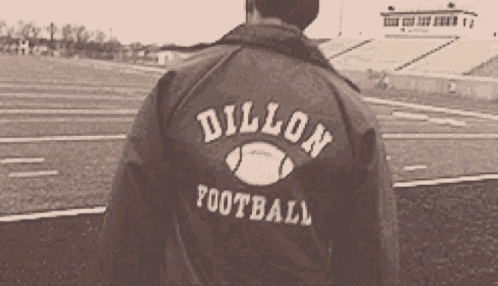 a man wearing a dillon football jacket stands on a football field