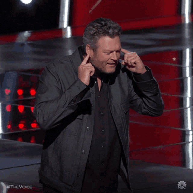 a man covering his ears with his hands on a stage with nbc written on the bottom right