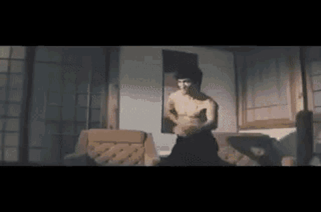 a shirtless man is standing in a room with a couch and a window .