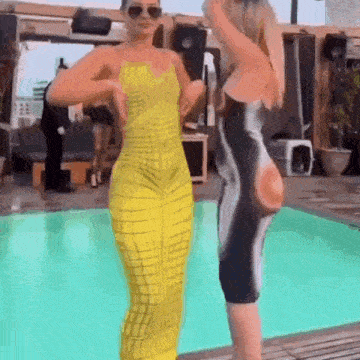 two women are standing next to each other in front of a pool . one of the women is wearing a yellow jumpsuit .