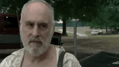 an older man with a beard and bald head is standing in a park looking at the camera .