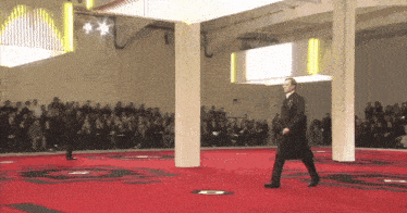a man in a black coat walks on a red carpet with the letter c on it