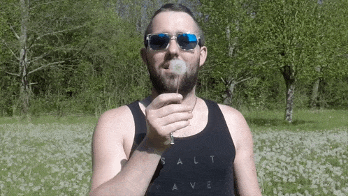a man blowing a dandelion wearing sunglasses and a black tank top that says salt wave