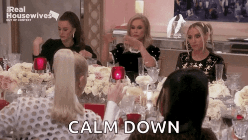 a group of women are sitting at a table with flowers and wine glasses and the word calm down is written on the screen .
