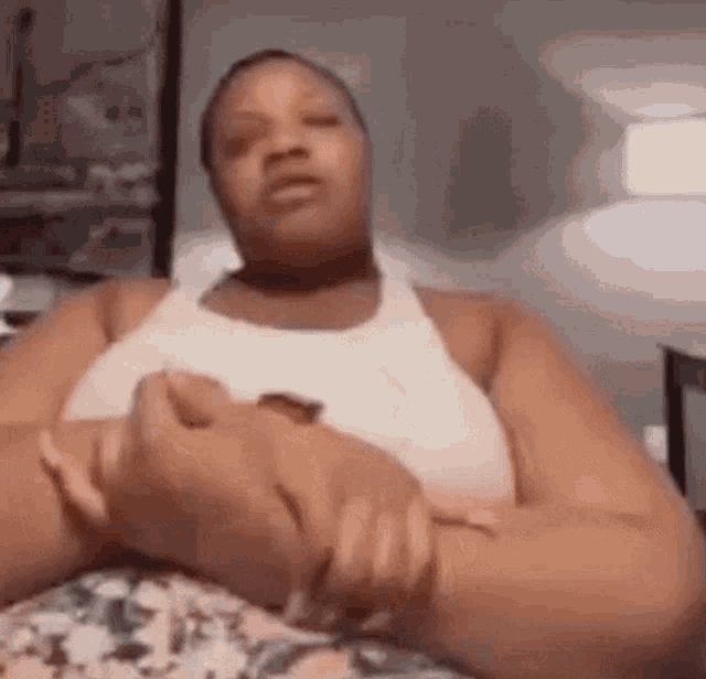 a woman in a white tank top is sitting on a bed with her arms crossed and looking at the camera .
