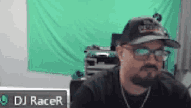 a man wearing a hat and glasses is sitting in front of a green screen and a sign that says dj racer .