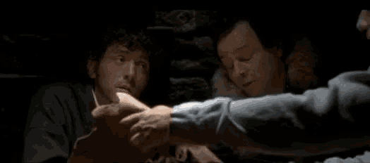 two men are standing next to each other in a dark room and one of them is reaching for something .