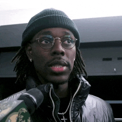 a man wearing a beanie and glasses holds a microphone