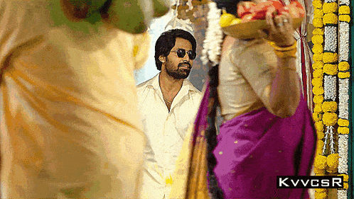a man wearing sunglasses stands in front of a woman in a purple saree
