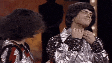 a man and a woman are standing next to each other . the man is wearing a silver jacket with sequins .