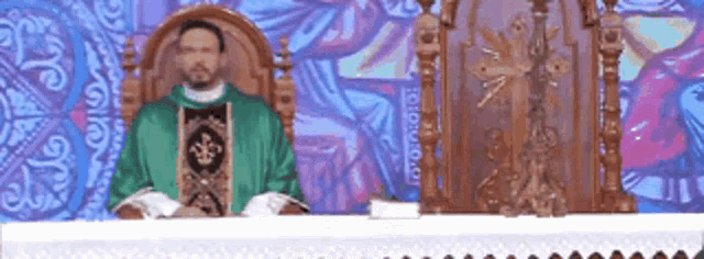 a priest in a green robe is sitting at a table in front of a painting