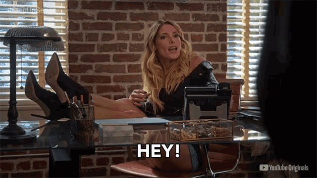 a woman sits at a desk with her legs crossed and says hey on the screen