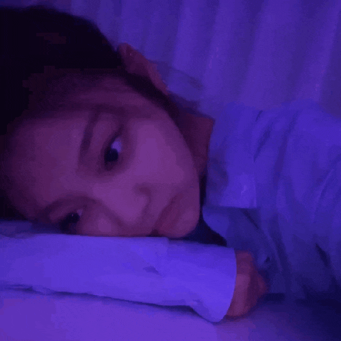 a woman is laying on a bed with her head on a pillow and a purple light behind her .