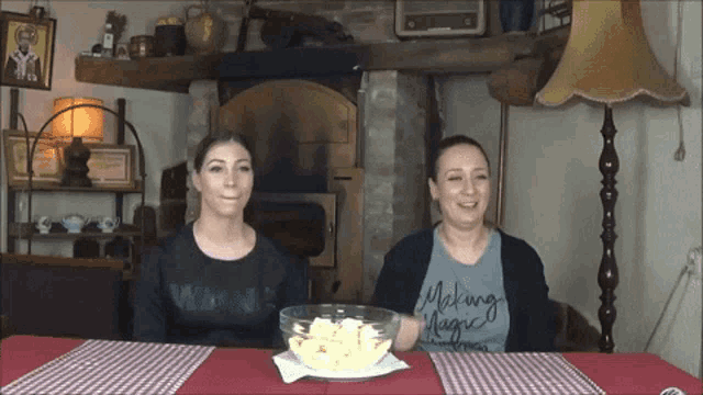 two women sit at a table with a bowl of food and one has a shirt that says making magic