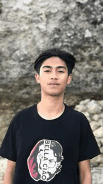 a young man in a black t-shirt is standing in front of a rocky cliff .