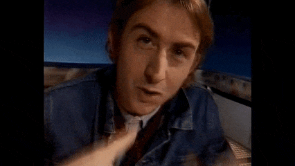 a man in a denim jacket and tie is making a funny face and pointing at the camera .