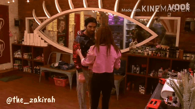 a woman in a pink jacket is hugging a man in front of a large eye shaped mirror made by kinemaster