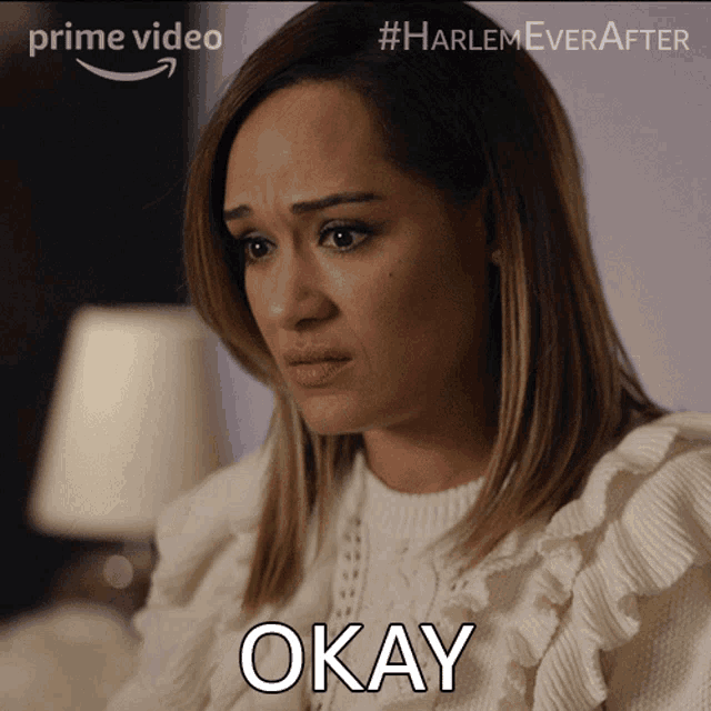 a woman in a white sweater says okay in front of a lamp