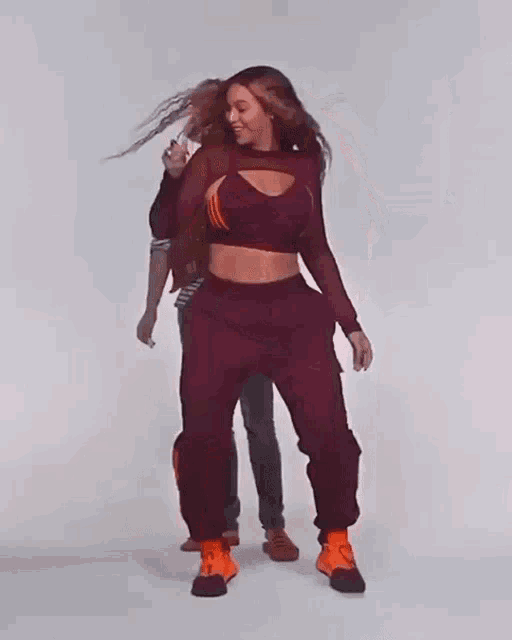 a woman in a red crop top and burgundy sweatpants is dancing while a man stands behind her .