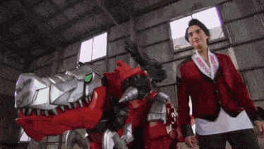 a man in a red jacket is standing next to a large toy dinosaur in a warehouse .