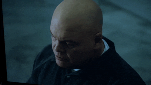 a bald man in a black jacket looks at the camera with a serious look on his face