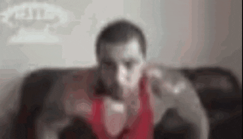 a blurry picture of a man in a red tank top sitting on a black couch .