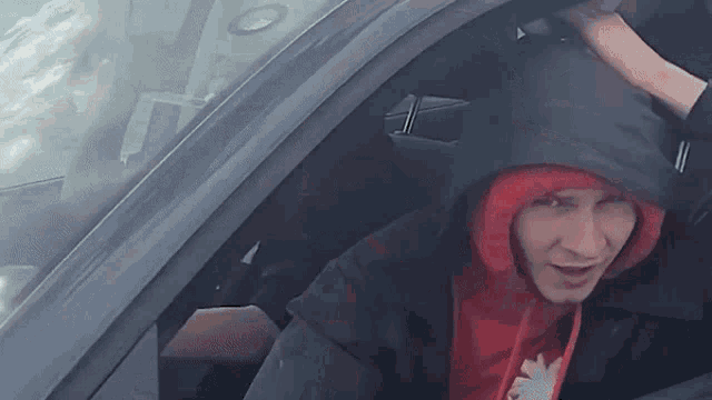 a man wearing a black jacket and a red hoodie is sitting in the driver 's seat of a car