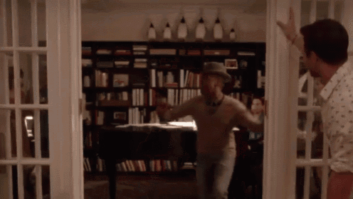 a man in a hat is dancing in front of a bookshelf in a living room