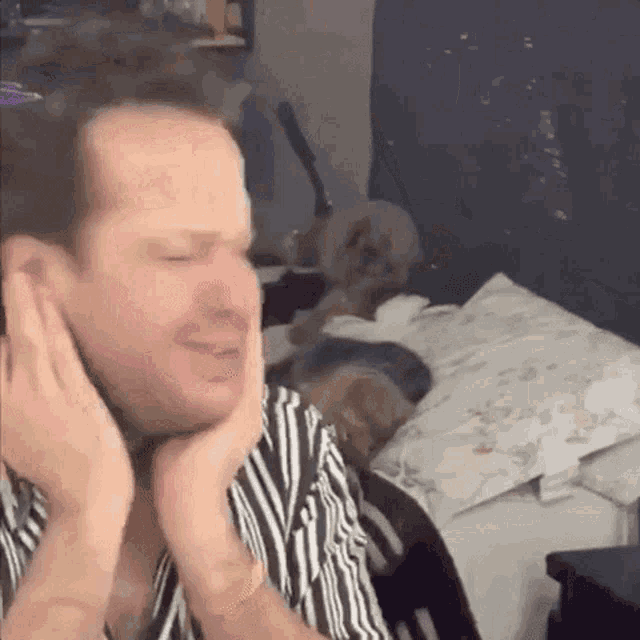 a man in a striped shirt is covering his ears with his hands in a bedroom .