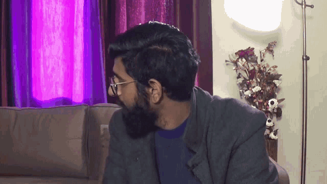 a man with glasses and a beard is sitting on a couch in a living room
