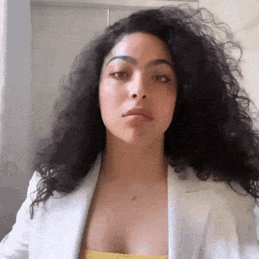 a woman with curly hair wearing a white jacket and a yellow top is looking at the camera .