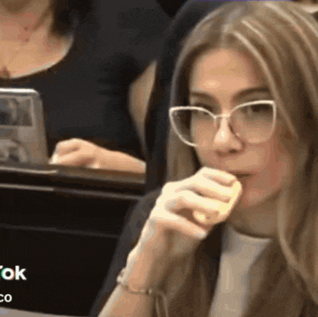a woman wearing glasses is sitting in a classroom and eating a piece of food .