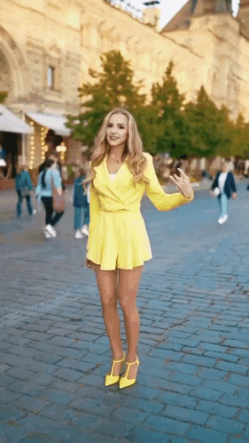 a woman in a yellow dress and high heels is standing on a brick sidewalk .