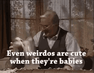 a man in a suit is sitting at a table holding a stuffed animal and talking about weird babies .