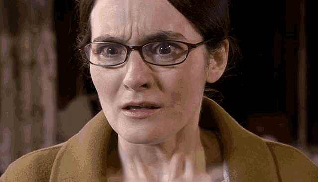 a woman wearing glasses and a brown coat is making a surprised face