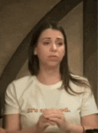 a woman in a white t-shirt is sitting down with her hands folded and looking at the camera .