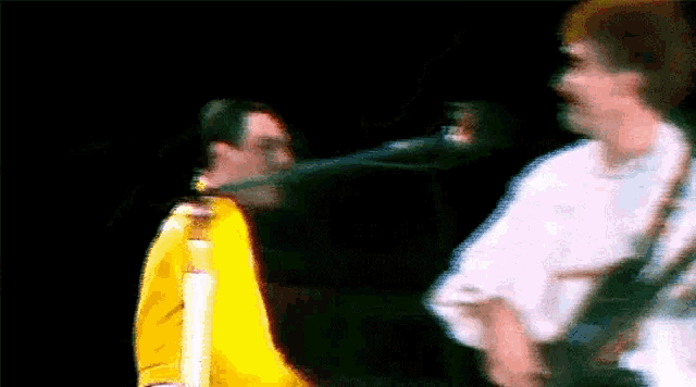 a man in a yellow jacket is standing next to a man in a white shirt on a stage playing a guitar .